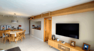 Sala de estar moderna com painel de TV em madeira, bancada de madeira com detalhes em ripado, vigas de madeira no teto, mesa de jantar em madeira clara com cadeiras, cozinha integrada e design contemporâneo em ambiente aberto; Creare Ambientes.