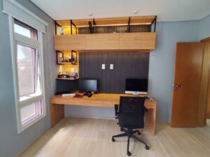 Escritório moderno e minimalista com parede cinza, dois monitores sobre mesa de madeira clara, cadeira preta giratória, prateleiras flutuantes em madeira com iluminação amarela acima, parede de painéis corrugados cinzento escuro, janela com vidros rosa à esquerda, porta de madeira à direita, piso em laminado claro e teto branco. Projeto produzido pela Creare Ambientes.